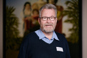 Image of Prof. Juckel wearing glasses and a sweater, standing in front of a painting.