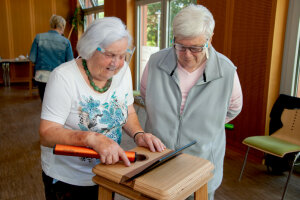 Two seniors are standing at a small wooden table with a tablet in it, handling an object similar to a baton. © Assunta Jaeger